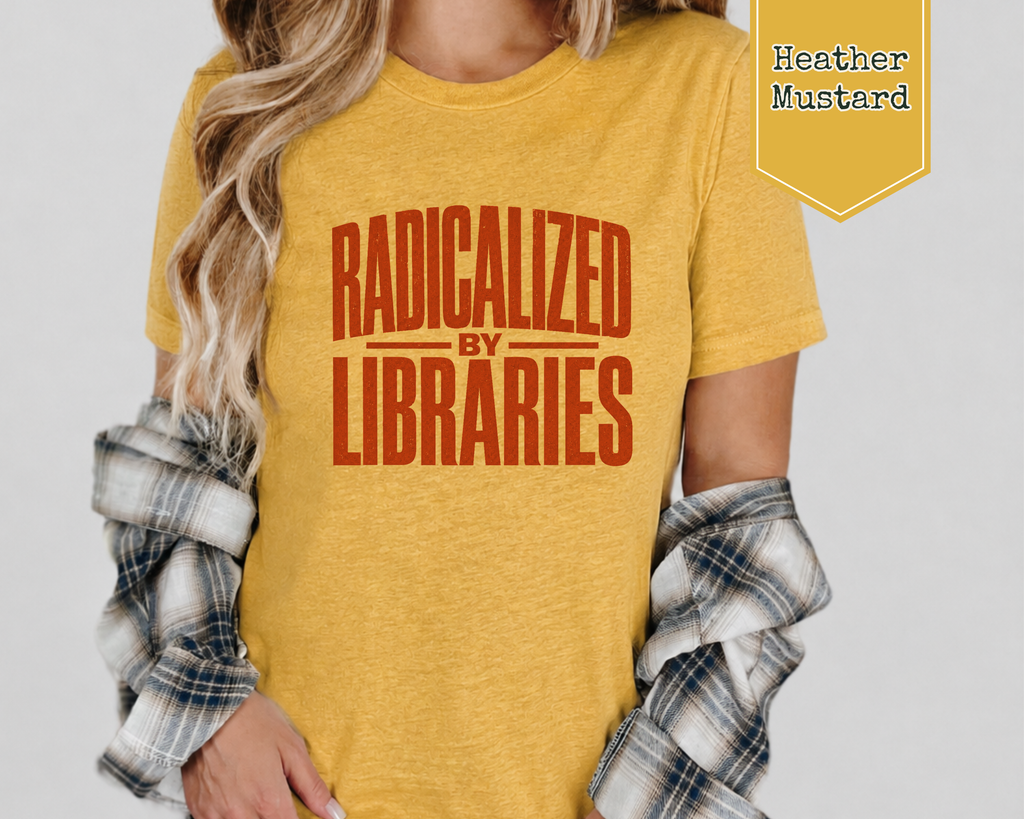 A woman wearing a heather mustard yellow graphic t-shirt with the text 'RADICALIZED BY LIBRARIES' in a distressed reddish-brown font. She has a plaid flannel shirt draped off her shoulders, and a yellow badge in the top right corner reads 'Heather Mustard'.