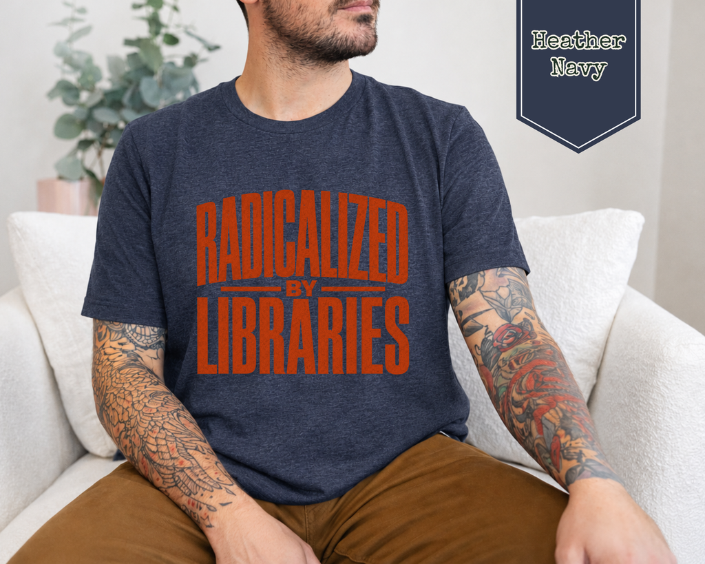 A man wearing a heather navy blue t-shirt with the words 'RADICALIZED BY LIBRARIES' printed in orange across the chest, sitting on a white couch. A badge in the top right corner reads 'Heather Navy'.