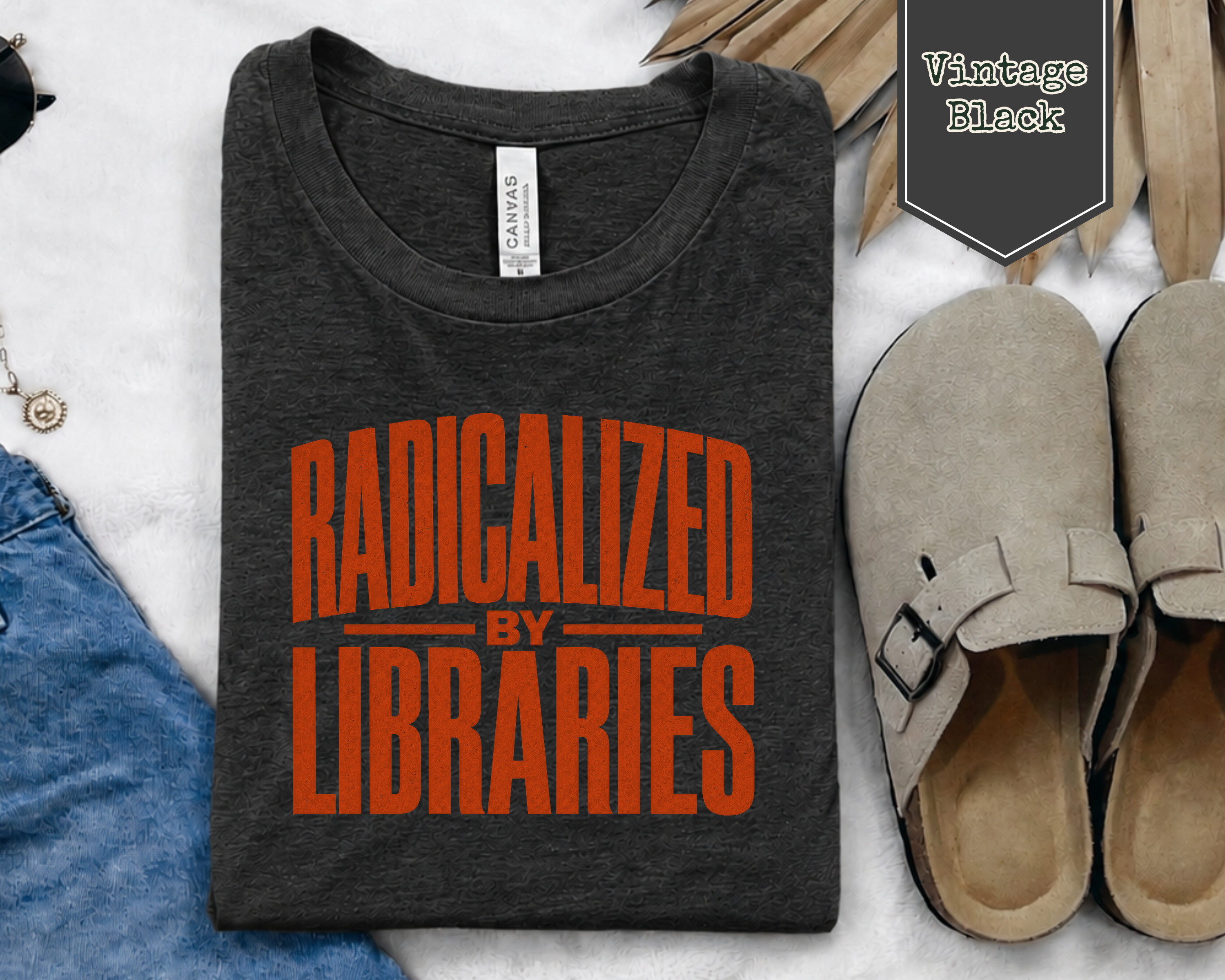 A folded dark charcoal heather graphic T-shirt with bold orange text reading 'RADICALIZED BY LIBRARIES' is shown in a styled flat lay on a white textured surface, alongside jeans, beige clogs, sunglasses, and a 'Vintage Black' label.