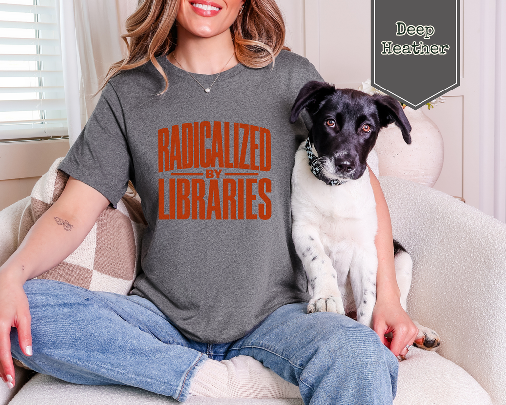 A woman sitting on a beige couch wearing a dark heather gray t-shirt with orange text that reads 'RADICALIZED BY LIBRARIES'. A black and white dog is sitting on her lap.