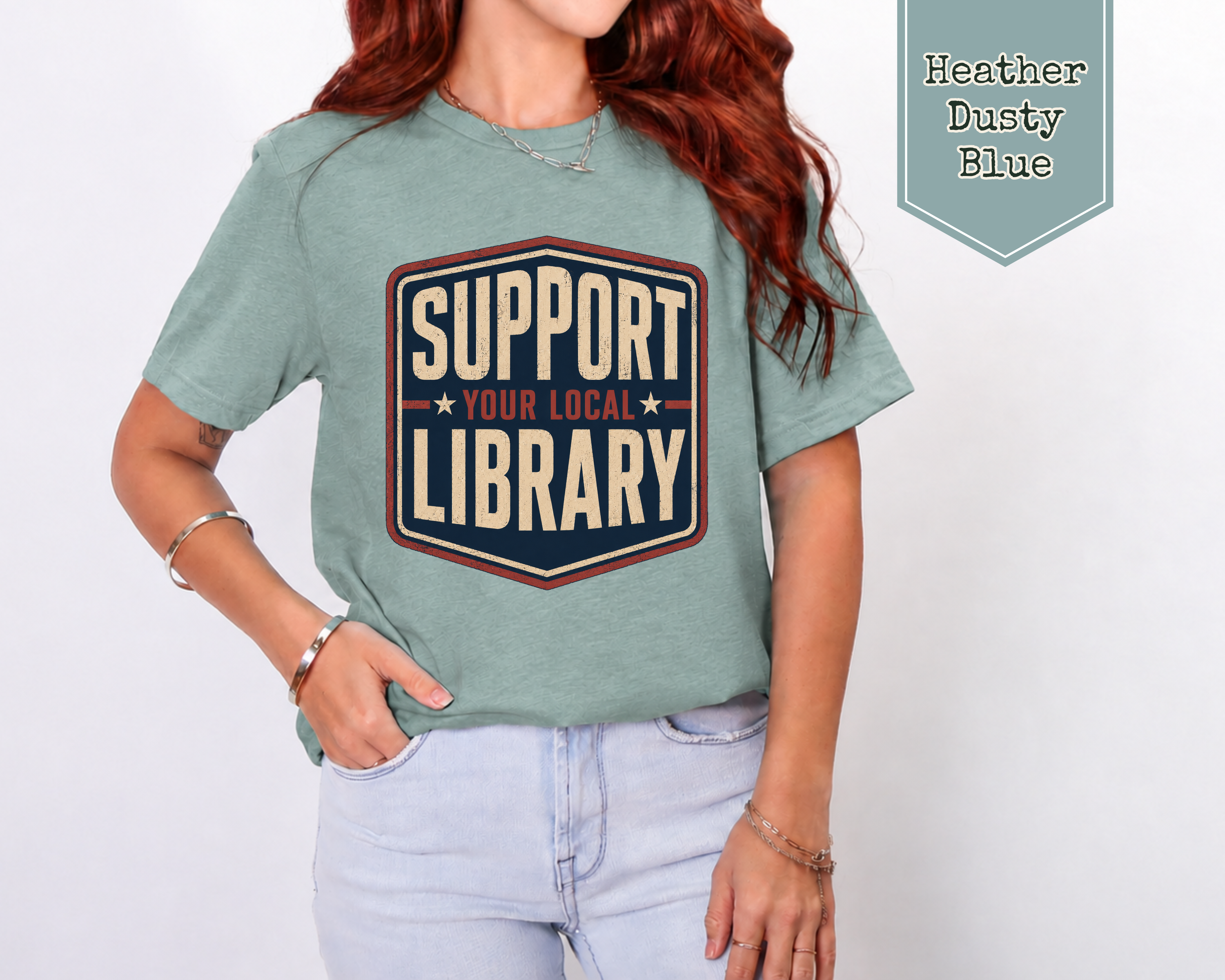 A woman wearing a heather dusty blue graphic t-shirt with a retro-style 'Support Your Local Library' print, paired with light-wash jeans. A teal badge in the top right corner reads 'Heather Dusty Blue'.