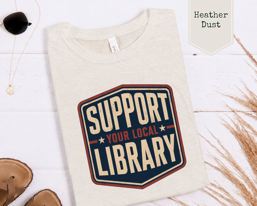 A folded Heather Dust heathered off-white t-shirt with a retro-style graphic that reads 'SUPPORT YOUR LOCAL LIBRARY' in cream and navy with red accents, styled on a white wood background with sunglasses, sandals, and dried grass.