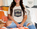 A woman sitting on a bed wearing a light heather dust graphic t-shirt that reads 'SUPPORT YOUR LOCAL LIBRARY'. The scene is decorated for autumn with pumpkins, a wicker basket, and a striped blanket.