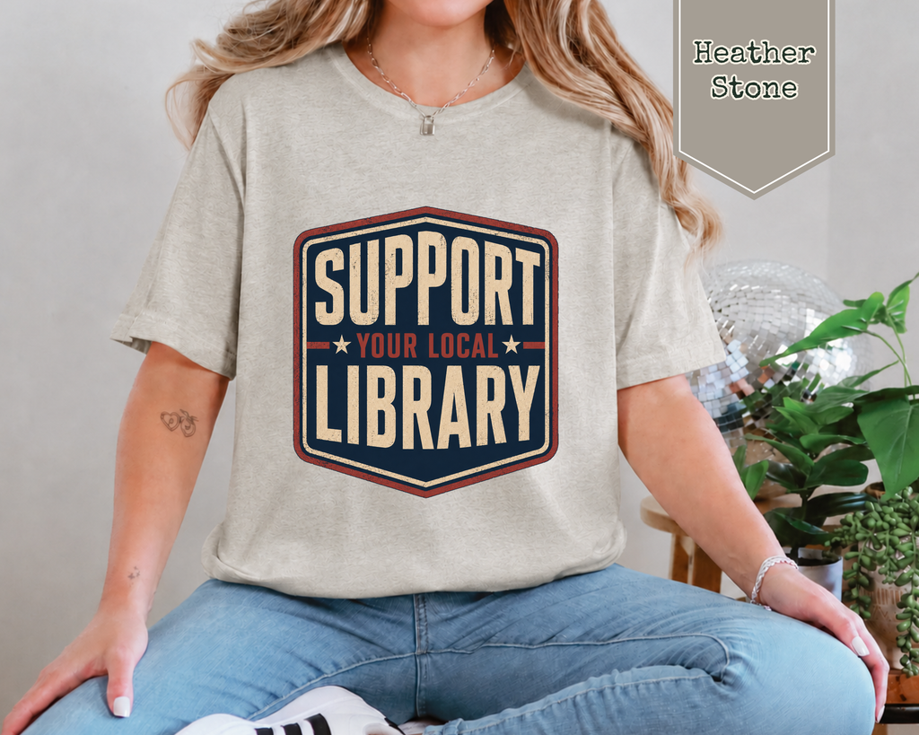 A woman wearing a light heather stone t-shirt with a retro-style graphic that reads 'SUPPORT YOUR LOCAL LIBRARY' in cream, navy, and red. She is sitting cross-legged in blue jeans, with a plant in the background and a 'Heather Stone' color label overlay in the top right corner.