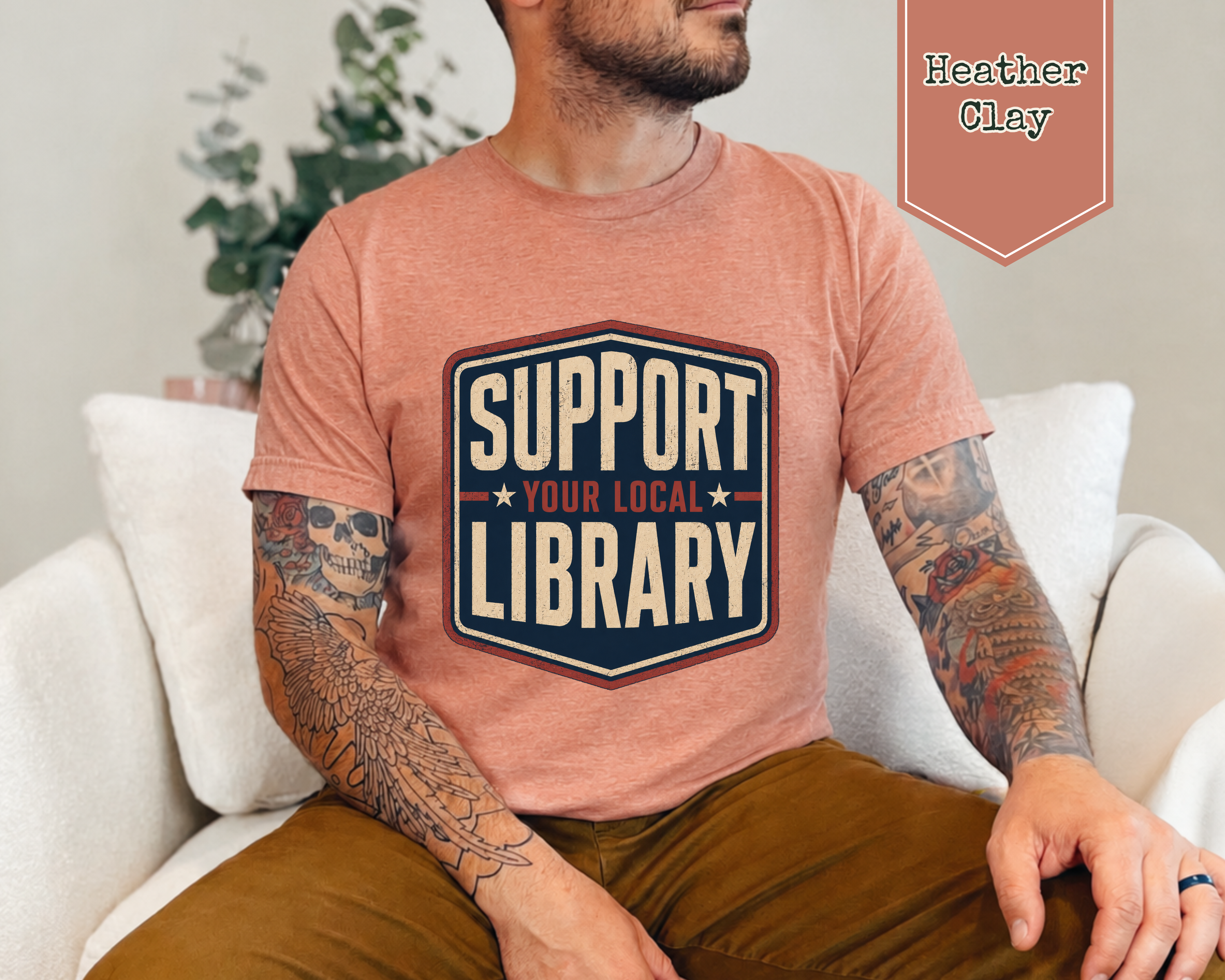 A man wearing a heather clay t-shirt with a 'Support Your Local Library' graphic, sitting on a white couch. A badge in the top right corner reads 'Heather Clay'.
