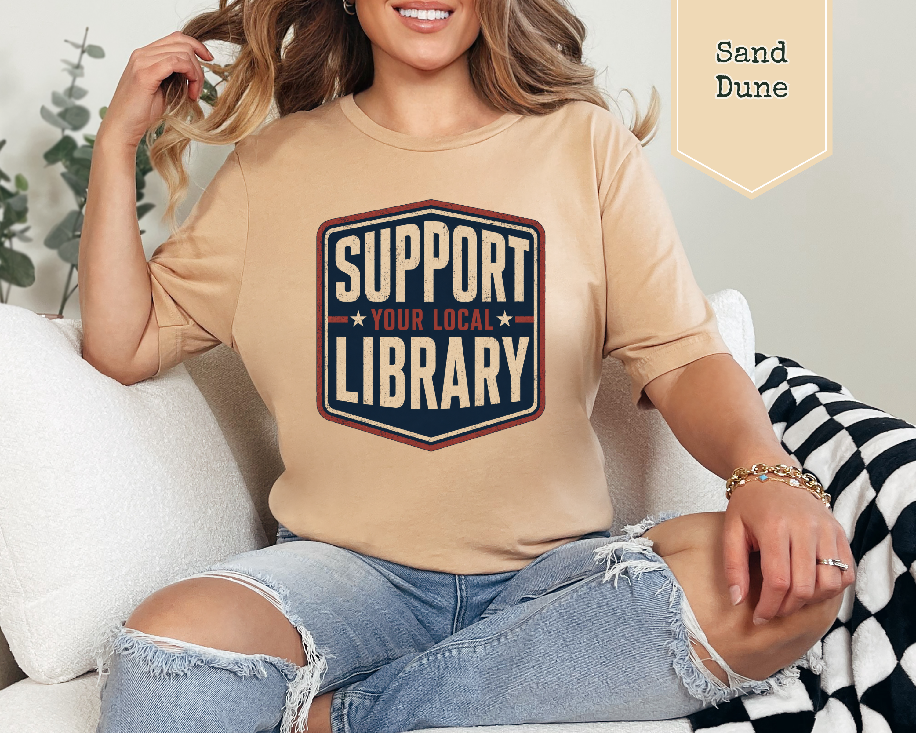 A woman sitting on a white couch wearing a beige graphic t-shirt with a navy and red shield design that reads 'SUPPORT YOUR LOCAL LIBRARY'. A beige banner in the top right corner reads 'Sand Dune'.