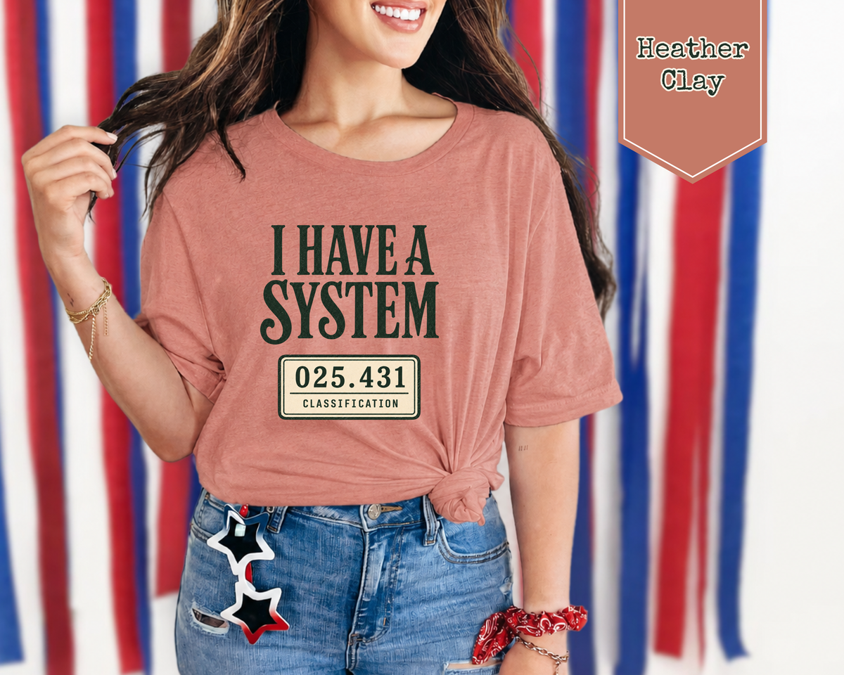 A woman wearing a heather clay pink graphic t-shirt with the text 'I HAVE A SYSTEM' and a license plate design, paired with blue jeans featuring a star patch. The background is a patriotic red, white, and blue striped banner, and a label in the top right reads 'Heather Clay'.