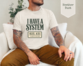A man sitting on a white couch wearing a light heather dust t-shirt with a dark green graphic that reads 'I HAVE A SYSTEM' and '025.431 CLASSIFICATION'.