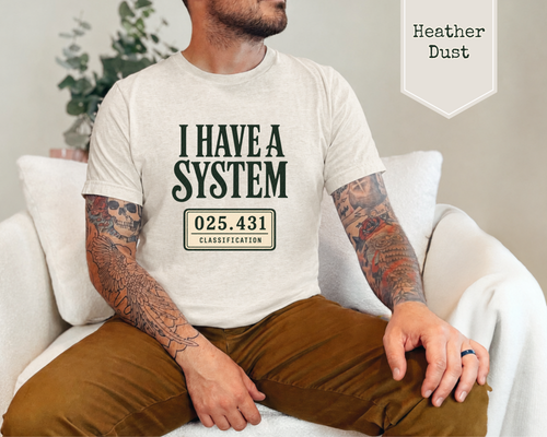 A man sitting on a white couch wearing a light heather dust t-shirt with a dark green graphic that reads 'I HAVE A SYSTEM' and '025.431 CLASSIFICATION'.
