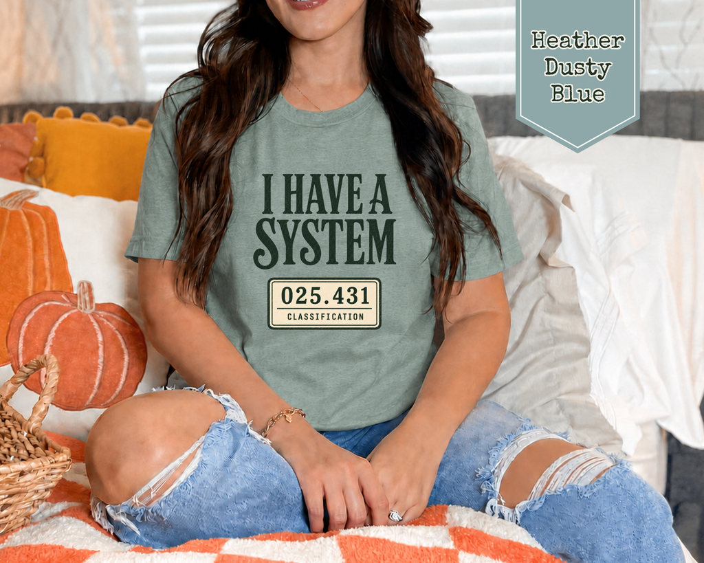 A woman sitting on a couch wearing a heather dusty blue graphic t-shirt with the text 'I HAVE A SYSTEM' and a license plate style design. The image includes a badge in the top right corner reading 'Heather Dusty Blue'.