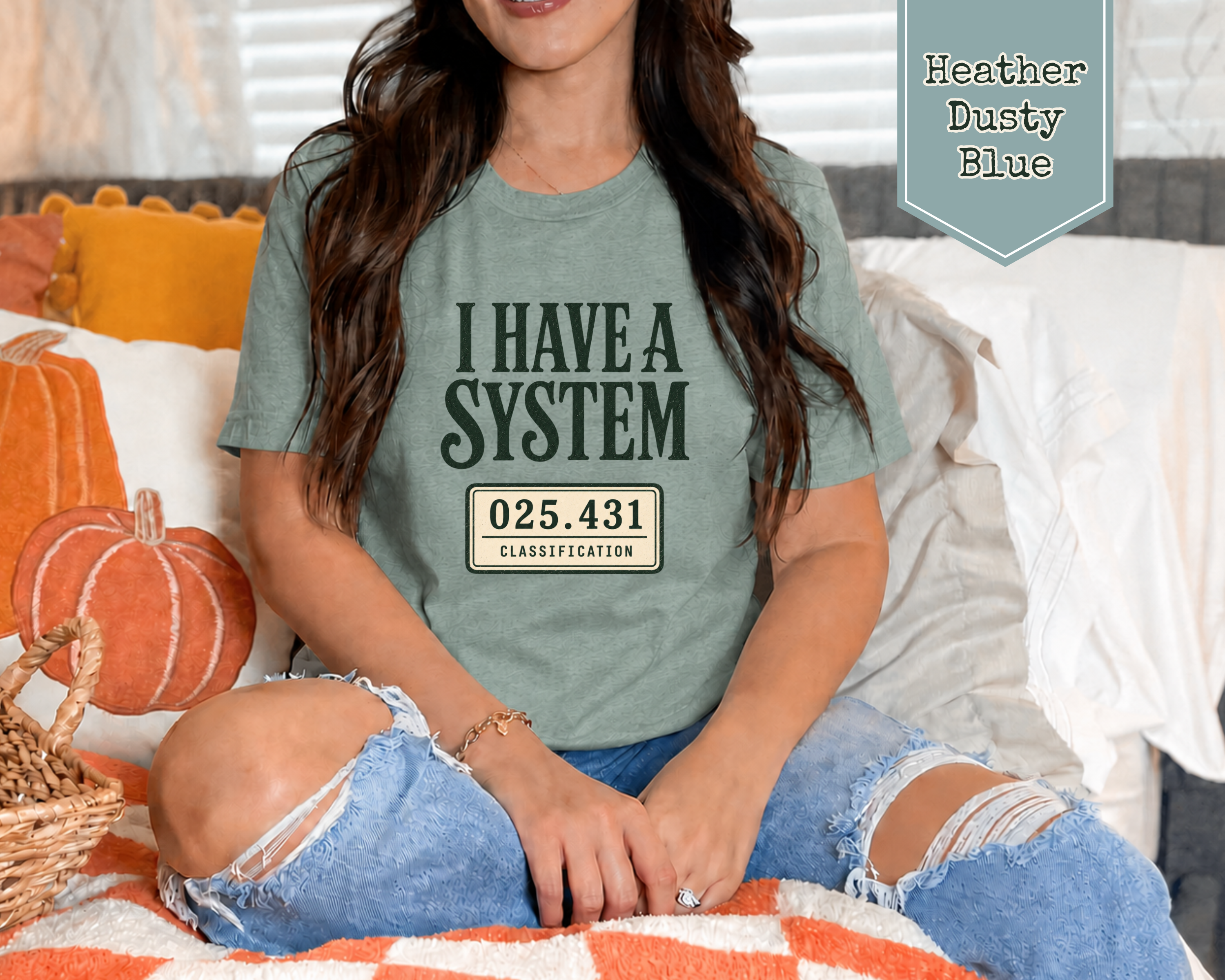 A woman sitting on a couch wearing a heather dusty blue graphic t-shirt with the text 'I HAVE A SYSTEM' and a license plate style design. The image includes a badge in the top right corner reading 'Heather Dusty Blue'.