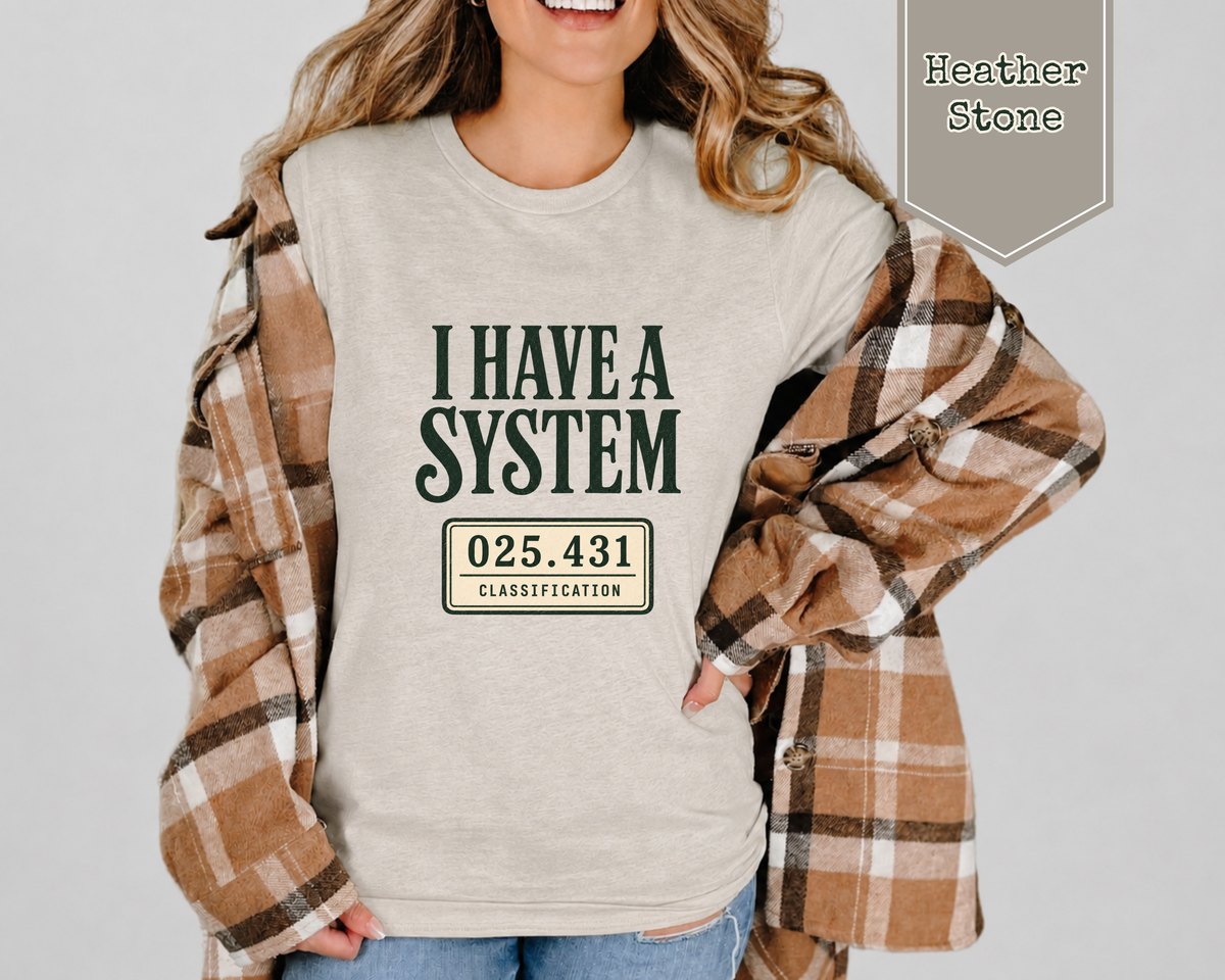 A woman wearing a light heather stone graphic t-shirt with the text 'I HAVE A SYSTEM 025.431 CLASSIFICATION' in dark green and beige.