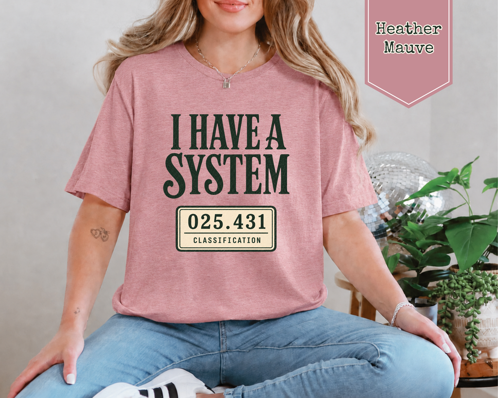 A woman wearing a heather mauve graphic t-shirt with the text 'I HAVE A SYSTEM' and a license plate design, paired with blue jeans. A pink badge in the top right corner reads 'Heather Mauve'.