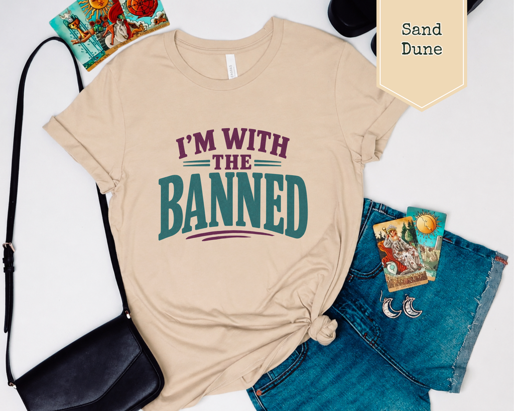 A shirt flat lay with a purse, jean shorts, sandals, and tarot cards. The t-shirt with purple and green phrase of I'm With The Banned.
