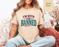 A woman sitting on a chair holding a mug. She is wearing a t-shirt with purple and green phrase of I'm With The Banned.