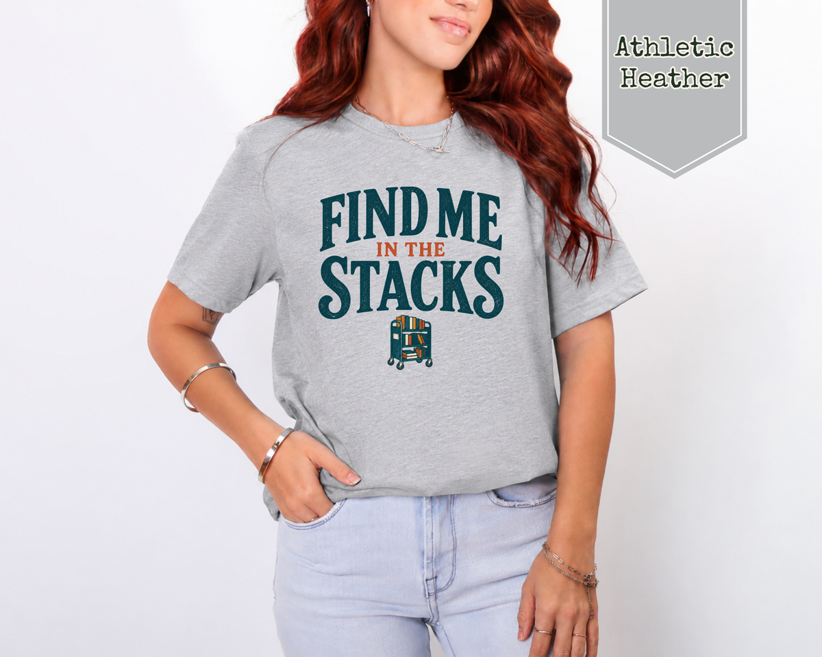 A woman wearing an athletic heather shirt. The t-shirt says Find Me In The Stacks with a small library book cart underneath.