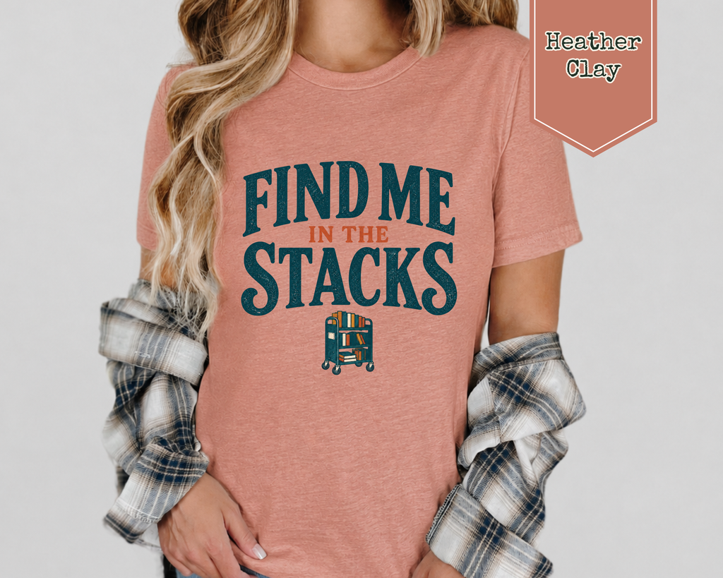 A woman wearing a heather clay t-shirt with a flannel long sleeve shirt. The t-shirt says Find Me In The Stacks with a small library book cart underneath.