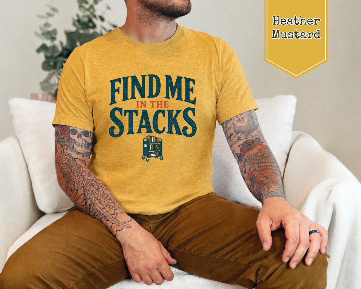 A man wearing a mustard yellow graphic t-shirt that reads 'FIND ME IN THE STACKS' with a small train illustration, sitting on a white couch. A yellow badge in the top right corner reads 'Heather Mustard'.