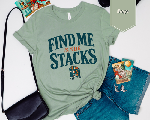 A sage green graphic t-shirt with the text 'FIND ME IN THE STACKS' and a small bus illustration, styled in a flat lay with denim shorts, a black crossbody bag, and tarot cards on a white background. A green label in the top right reads 'Sage'.