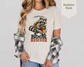 A woman wearing a light heather dust graphic t-shirt with a raccoon holding books and the words 'CERTIFIED BOOK RESCUEER'. She has a plaid shirt draped off her shoulders and is wearing blue jeans.