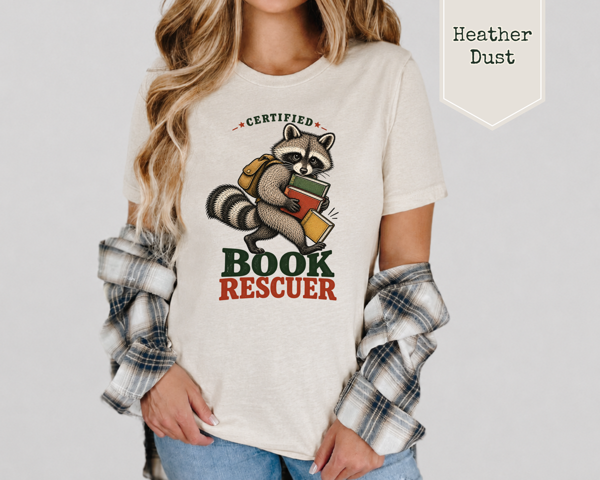 A woman wearing a light heather dust graphic t-shirt with a raccoon holding books and the words 'CERTIFIED BOOK RESCUEER'. She has a plaid shirt draped off her shoulders and is wearing blue jeans.
