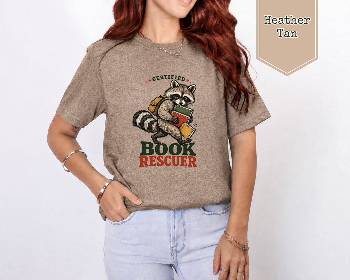 A woman wearing a heather tan graphic t-shirt with a raccoon holding books and the words 'CERTIFIED BOOK RESCUEER'. The image includes a 'Heather Tan' color label overlay in the top right corner.