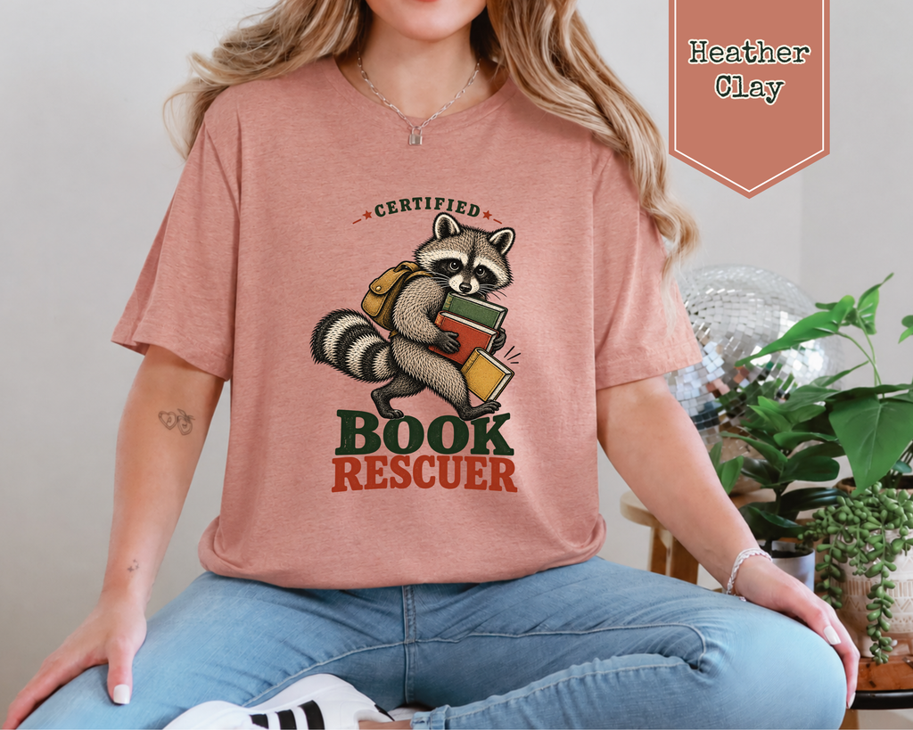 A woman wearing a heather clay pink graphic t-shirt with a cartoon raccoon holding books and the words 'CERTIFIED BOOK RESCUER'. The image includes a color label 'Heather Clay' in the top right corner.