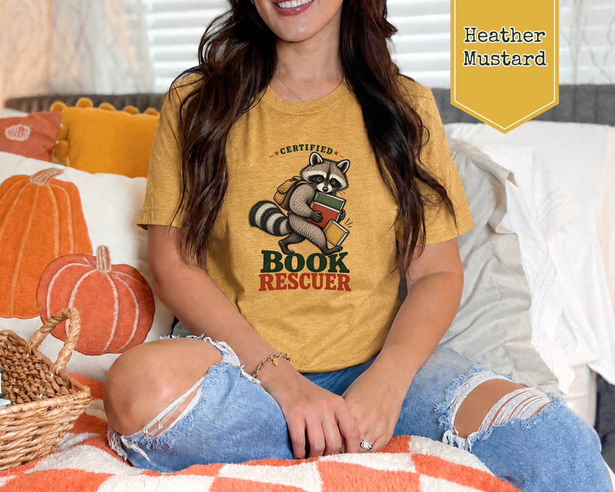 A woman wearing a mustard yellow graphic t-shirt with a raccoon holding a book and the words 'CERTIFIED BOOK RESCUEER'. She is sitting on a bed with autumn-themed pillows and pumpkins.