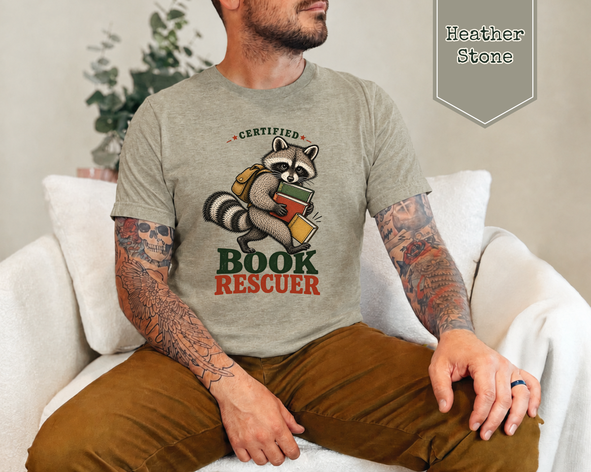 A man wearing a heather gray graphic t-shirt with a cartoon raccoon holding a book and the words 'CERTIFIED BOOK RESCUER' in green and orange. He is sitting on a white couch, and a badge in the top right corner reads 'Heather Stone'.