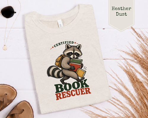 A folded cream-colored t-shirt with a graphic of a raccoon carrying books, labeled 'CERTIFIED BOOK RESCUER'. The t-shirt is displayed in a flat-lay arrangement on a white wood background with decorative props including sunglasses, a necklace, sandals, and dried grass.