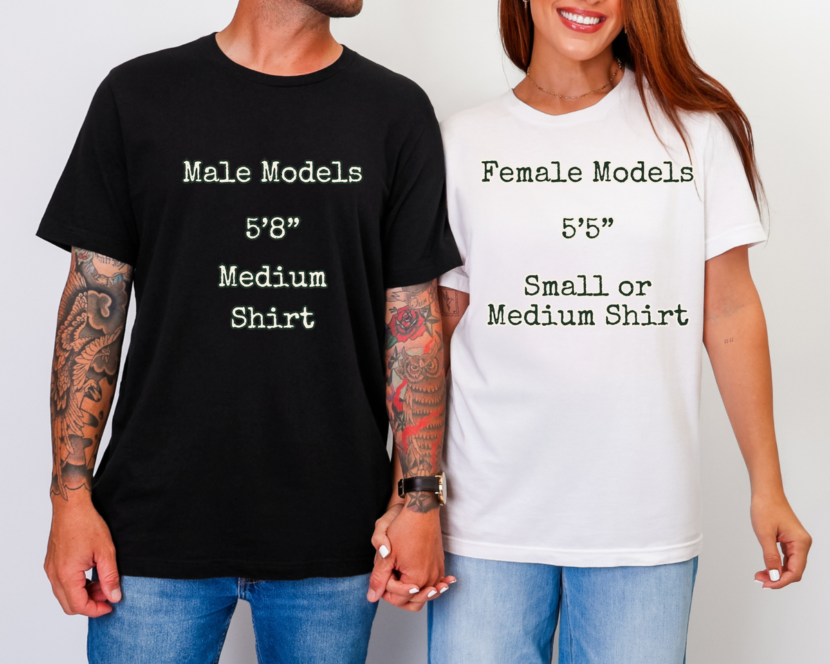 A man and a woman standing side by side holding hands, wearing matching graphic t-shirts. The man on the left wears a black t-shirt with white text reading 'Male Models 5'8" Medium Shirt', and the woman on the right wears a white t-shirt with black text reading 'Female Models 5'5" Small or Medium Shirt'.