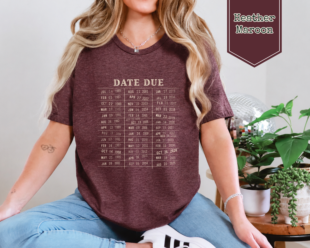 A woman wearing a heather maroon graphic t-shirt with a beige 'DATE DUE' calendar print, paired with blue jeans, sitting in a home setting with plants in the background.