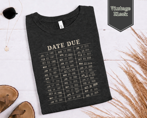 A folded black t-shirt with a beige 'DATE DUE' calendar print, styled in a flat-lay on a white wood background with sunglasses, sandals, and dried grass.