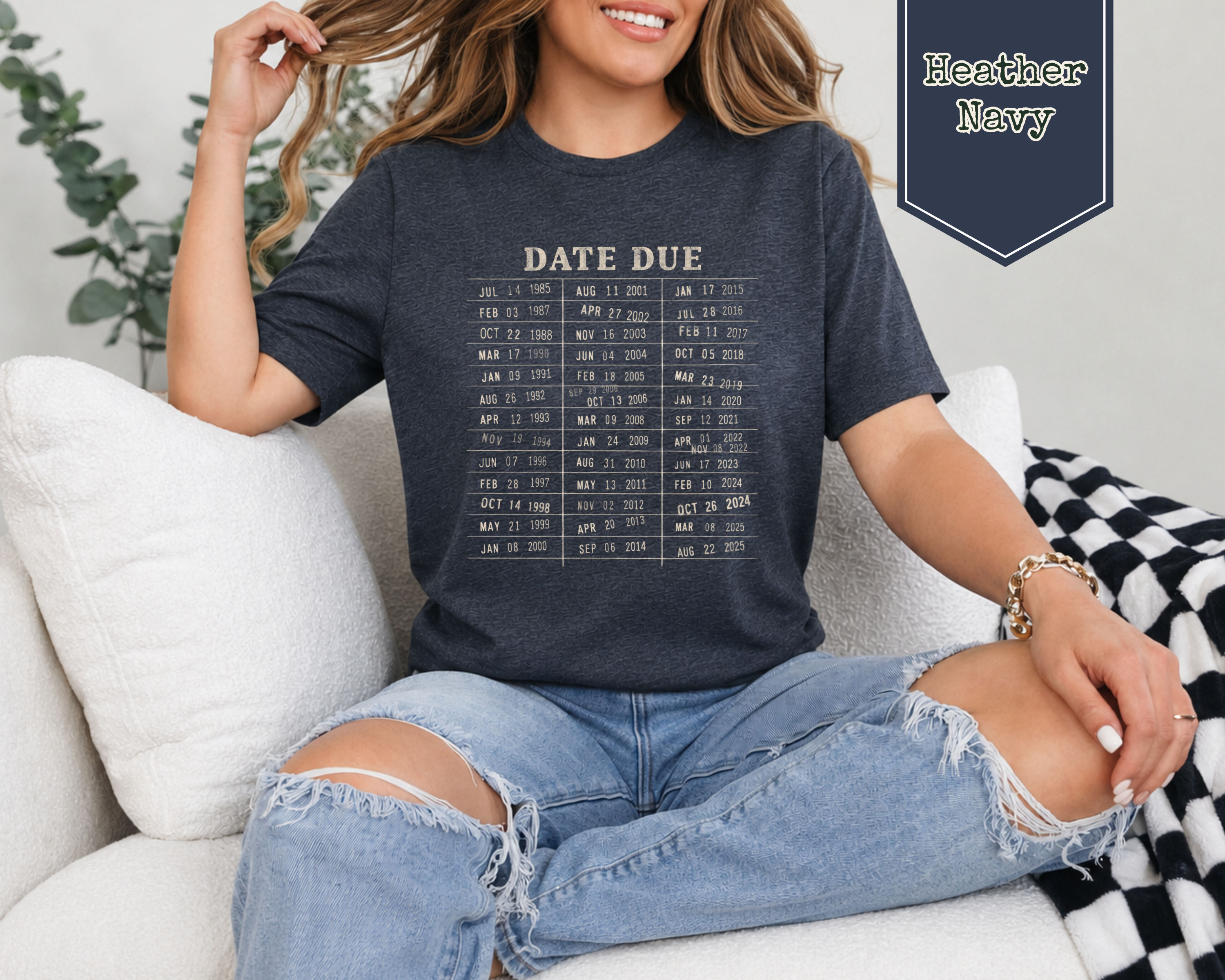 A woman sitting on a white couch wearing a dark heather navy graphic t-shirt with a calendar-style 'DATE DUE' print. She is wearing ripped light-wash jeans, and a navy label in the top right reads 'Heather Navy'.
