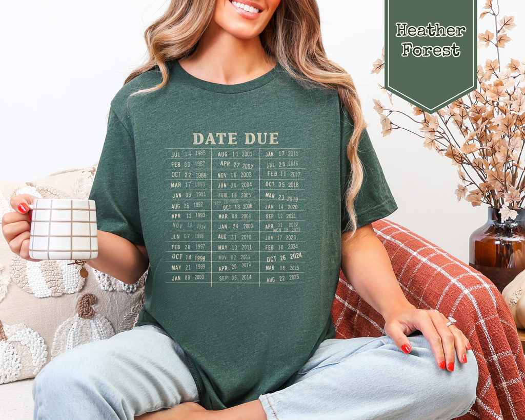 A woman sitting on a couch wearing a dark forest green t-shirt with a beige 'DATE DUE' calendar graphic. She is holding a mug, with a plaid throw blanket and a decorative banner in the background.