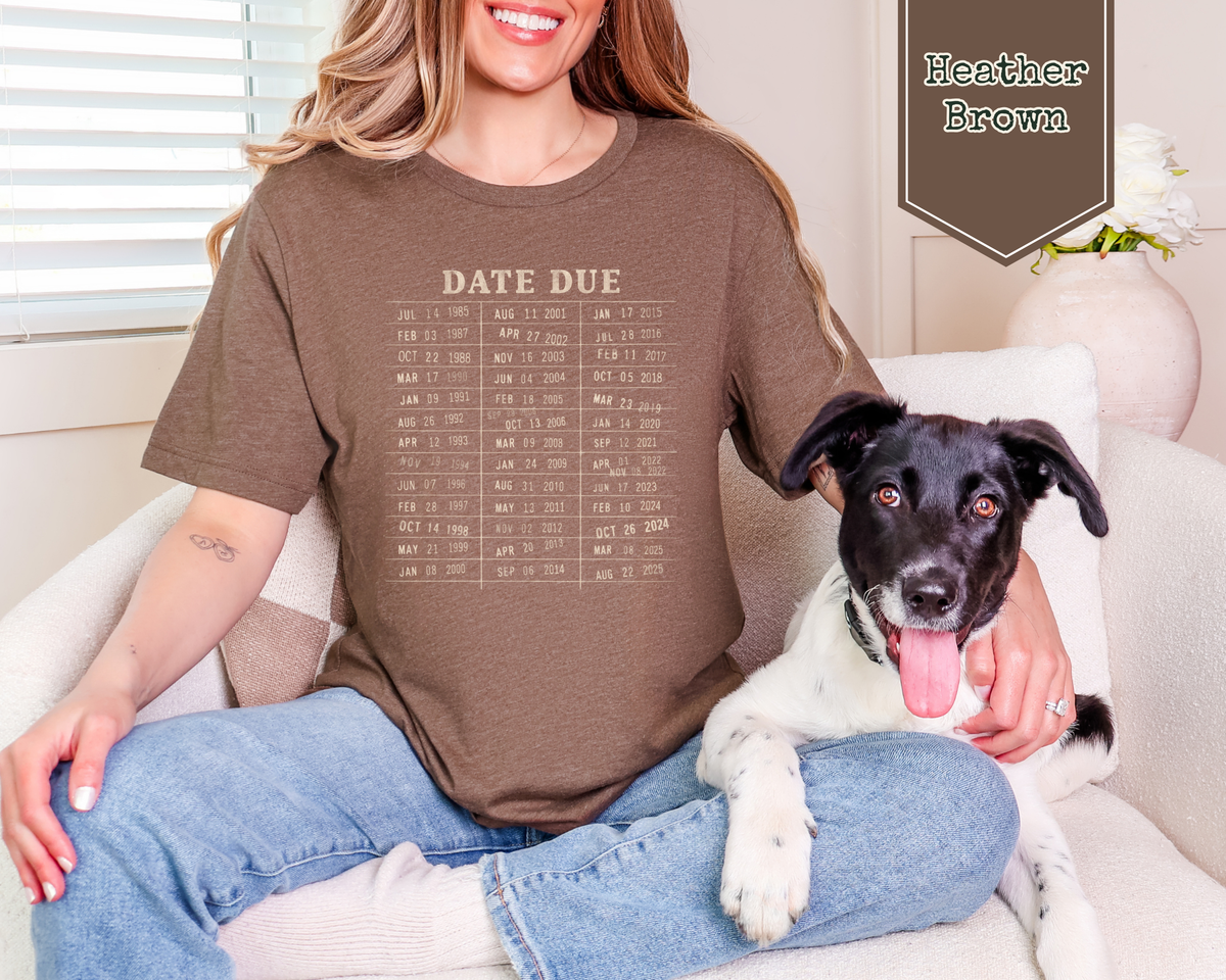 A woman sitting on a white couch wearing a brown graphic t-shirt with a 'DATE DUE' calendar print, holding a black and white dog. A brown badge in the top right corner reads 'Heather Brown'.