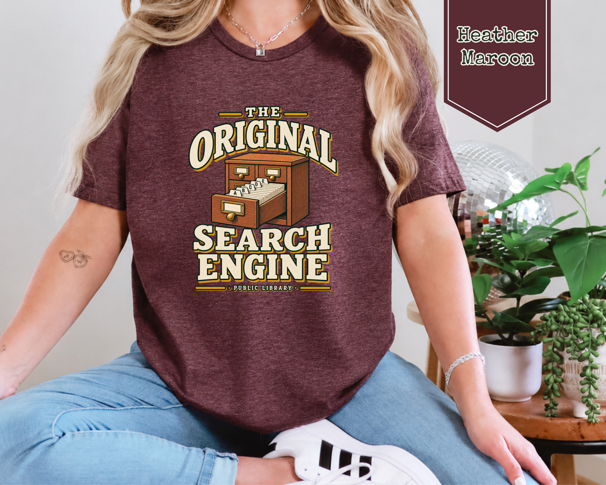 A woman wearing a heather maroon graphic t-shirt with a vintage-style 'The Original Search Engine' library logo. The image includes a color swatch label for 'Heather Maroon' and a background of indoor plants and a table.
