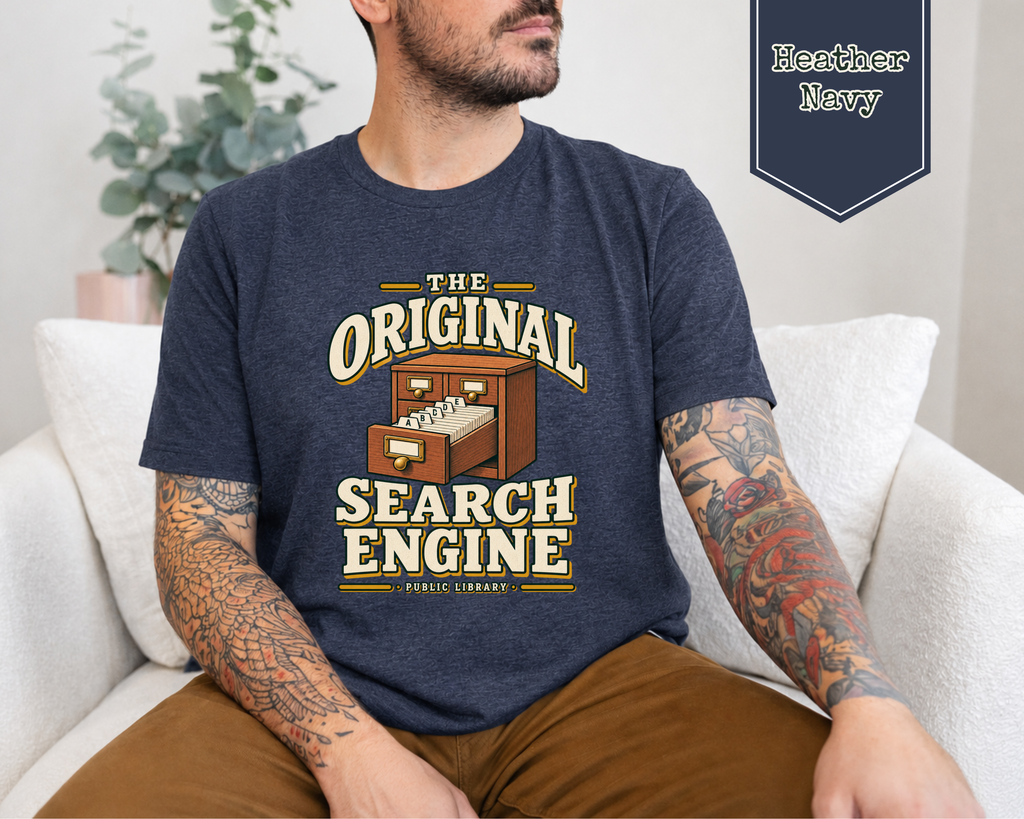 A man wearing a navy blue graphic t-shirt with a vintage-style 'The Original Search Engine' print, sitting on a white couch. A badge in the top right corner reads 'Heather Navy'.