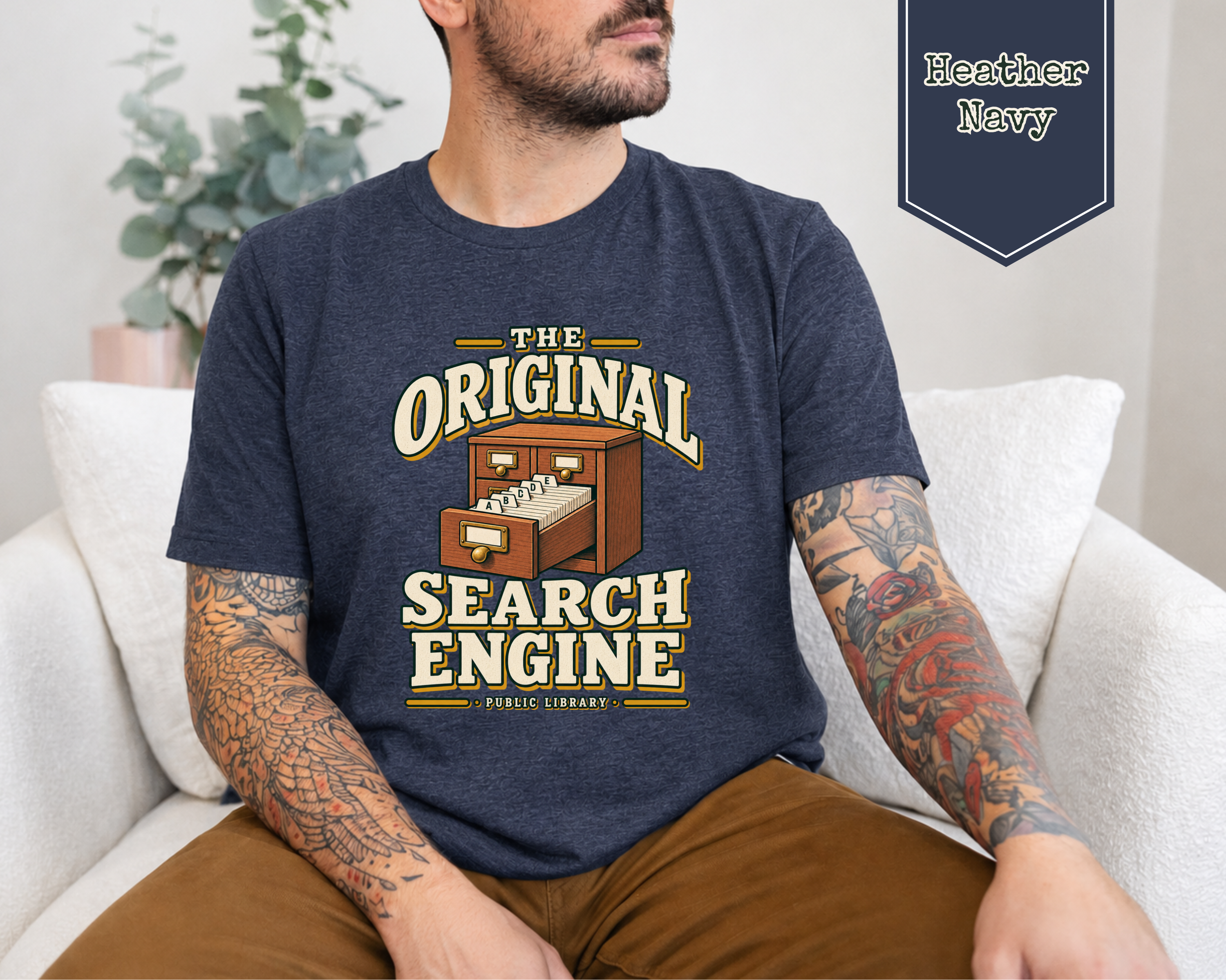 A man wearing a navy blue graphic t-shirt with a vintage-style 'The Original Search Engine' print, sitting on a white couch. A badge in the top right corner reads 'Heather Navy'.