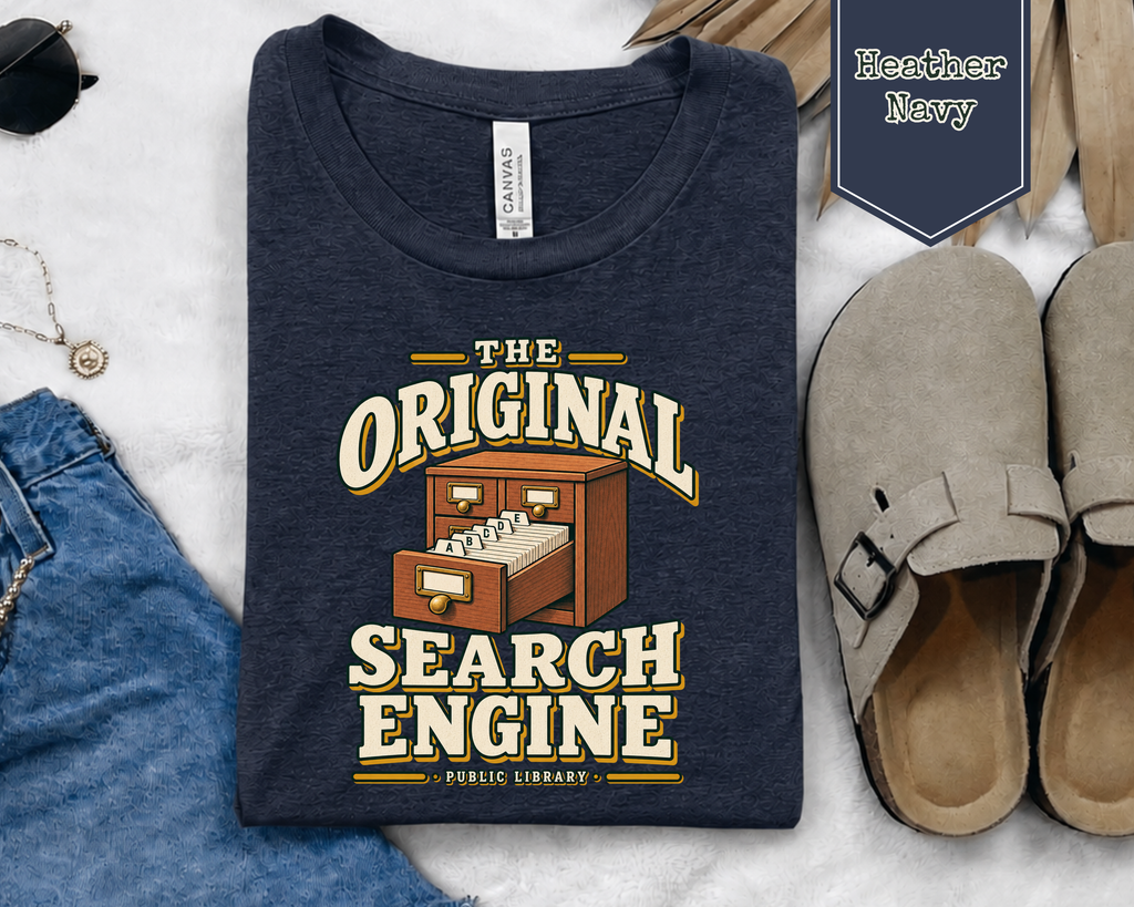 A navy blue t-shirt with a retro graphic reading 'THE ORIGINAL SEARCH ENGINE PUBLIC LIBRARY' in cream and yellow, styled in a flat lay with jeans, sunglasses, a cardholder, and clogs on a white marble background.