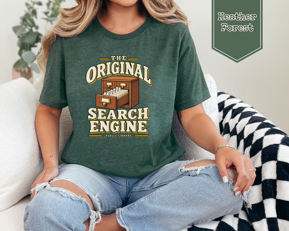 A woman sitting on a couch wearing a dark forest green graphic t-shirt with a retro computer graphic and the text 'THE ORIGINAL SEARCH ENGINE'. A badge in the top right corner reads 'Heather Forest'.