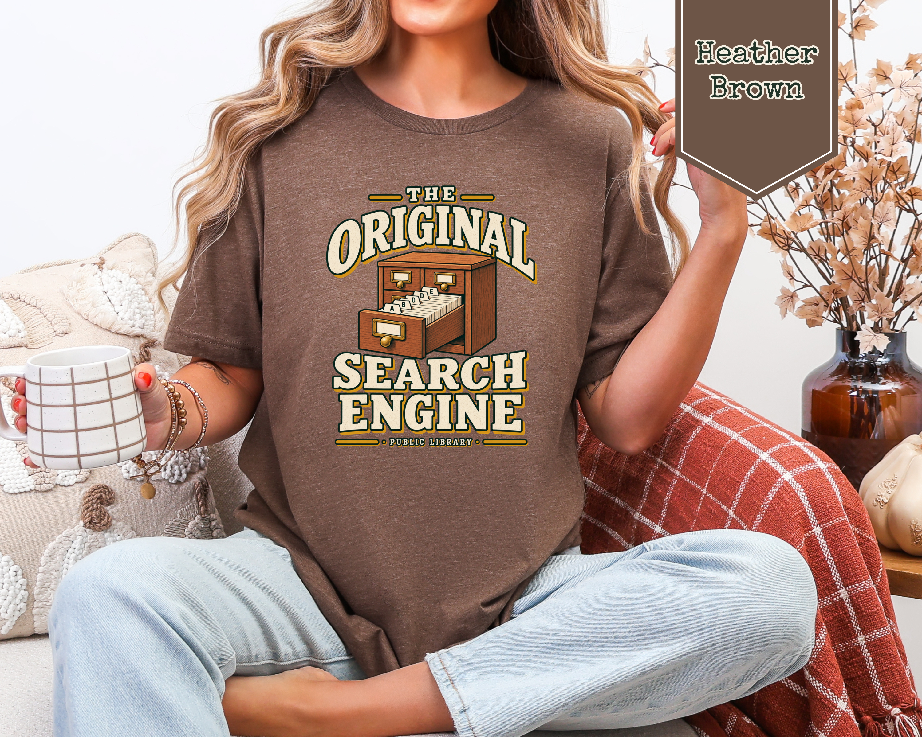 A woman wearing a brown graphic t-shirt with the text 'THE ORIGINAL SEARCH ENGINE' and a computer graphic, sitting in a cozy room setting with a mug and a plaid blanket.