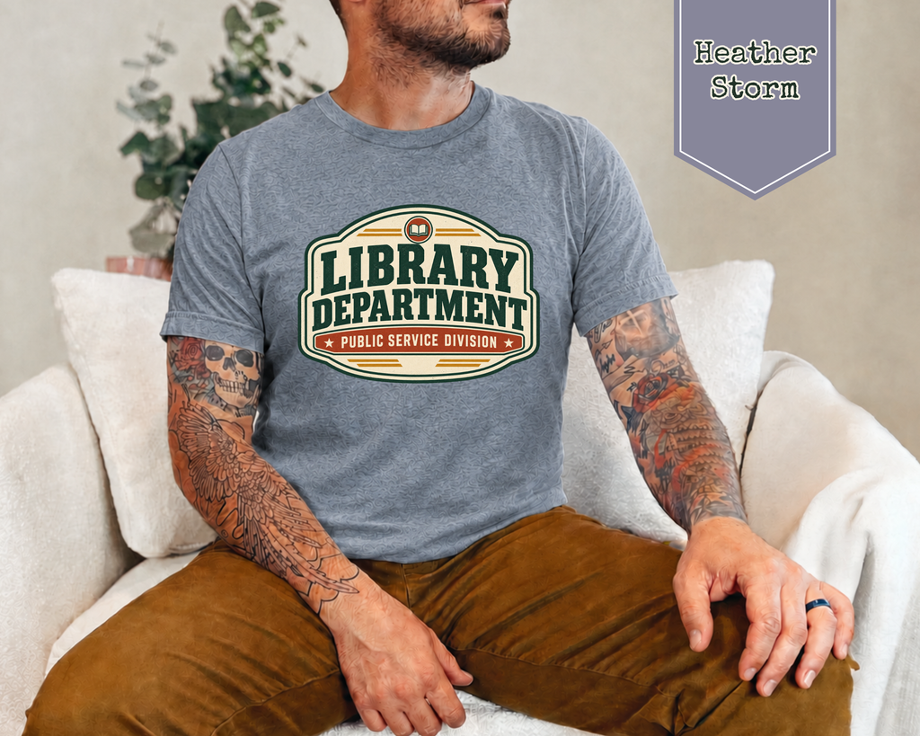 A man wearing a heather gray graphic t-shirt with a 'Library Department' print, sitting on a white couch. The image includes a purple badge in the top right corner labeled 'Heather Storm'.
