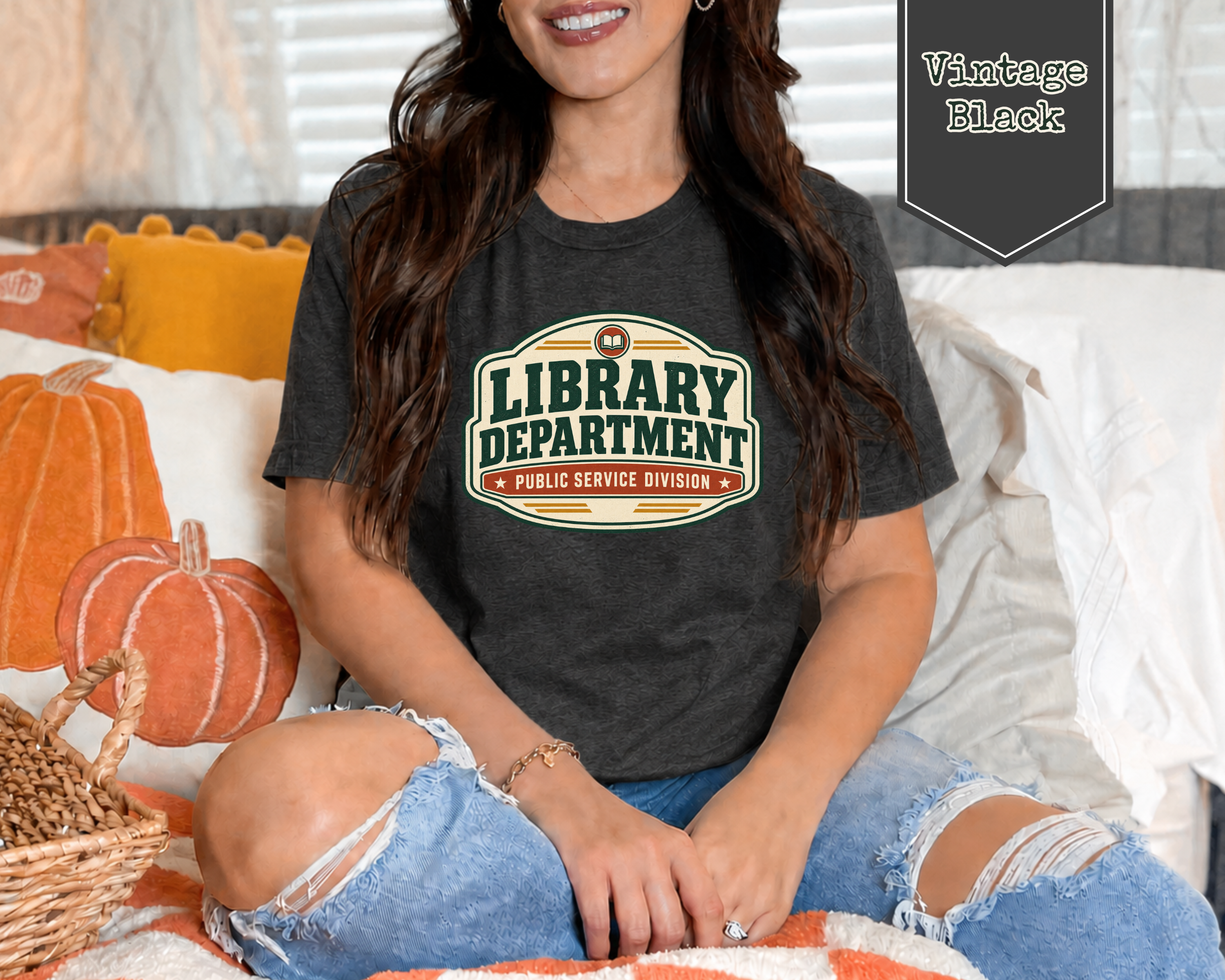 A woman sitting on a bed wearing a dark charcoal gray graphic t-shirt that reads 'LIBRARY DEPARTMENT PUBLIC SERVICE DIVISION' in a vintage-style badge. The image has a 'Vintage Black' label overlay in the top right corner.