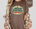 A woman wearing a heather brown graphic t-shirt with a 'Library Department' print, styled with a beige and brown plaid flannel shirt draped off her shoulders, against a light gray background.