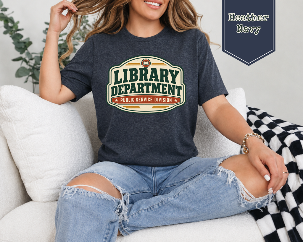 A woman sitting on a white couch wearing a heather navy graphic t-shirt with a retro 'Library Department Public Service Division' design. She is wearing distressed light blue jeans, and a navy blue badge overlay in the top right reads 'Heather Navy'.