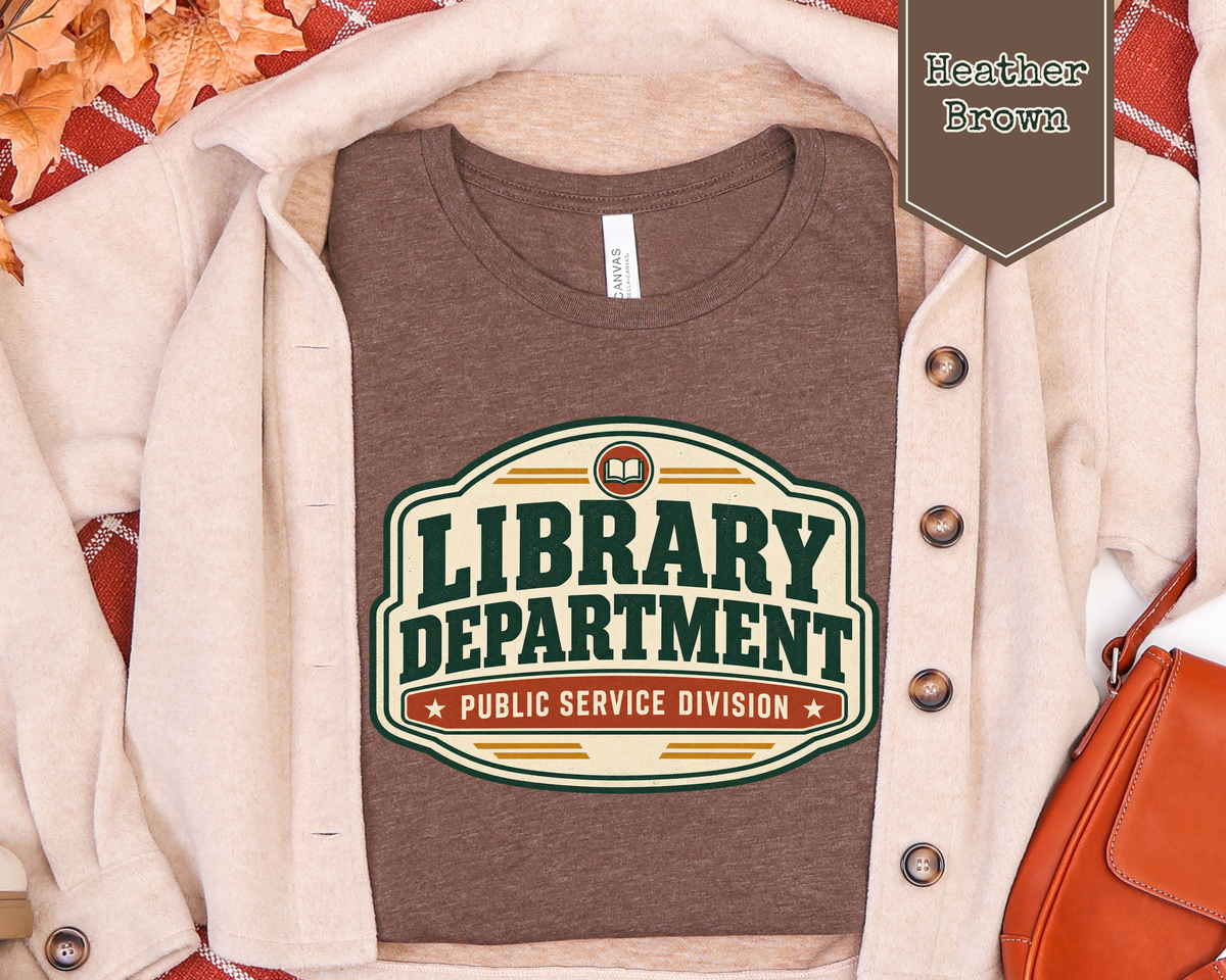 A brown heather graphic T-shirt styled in a flat lay with a cream cardigan, autumn leaves, and a brown bag. The shirt features a retro-style graphic that reads 'LIBRARY DEPARTMENT PUBLIC SERVICE DIVISION'.