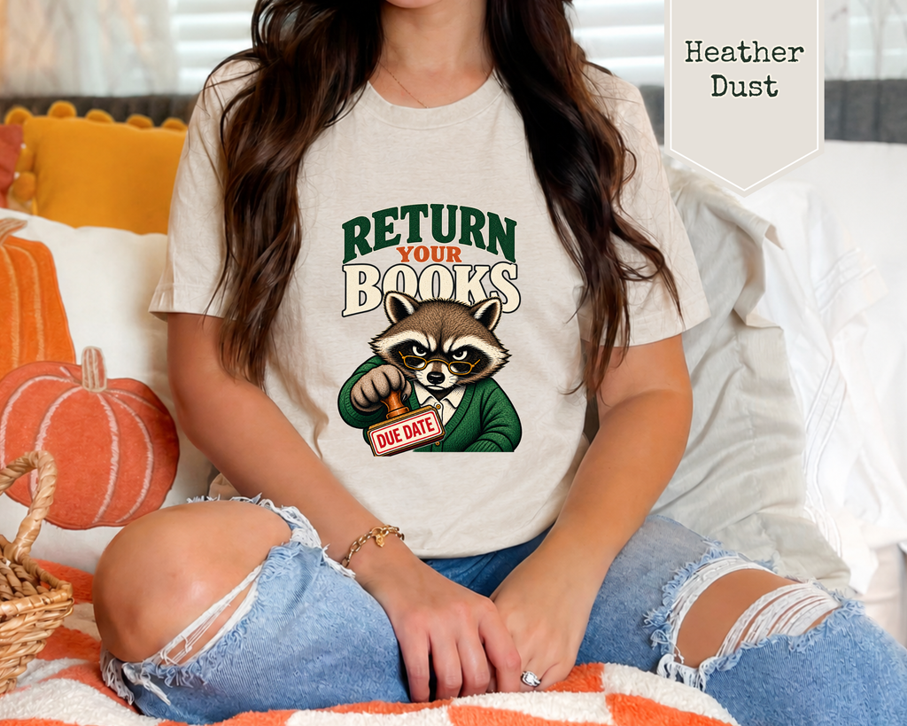 A woman wearing a light heather dust graphic t-shirt with a raccoon holding a red 'Date' sign and the text 'RETURN YOUR BOOKS'. She is sitting on a cozy bed with orange pumpkins and white pillows.