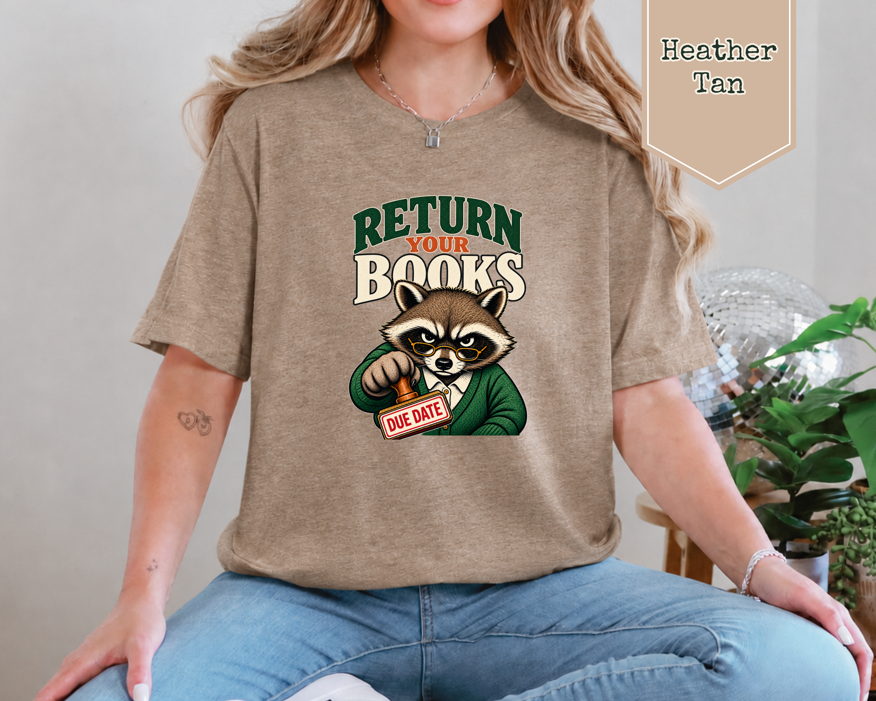 A woman wearing a heather tan graphic t-shirt with a raccoon holding a 'DUE DATE' sign and the text 'RETURN YOUR BOOKS'. A 'Heather Tan' color label is visible in the top right corner.