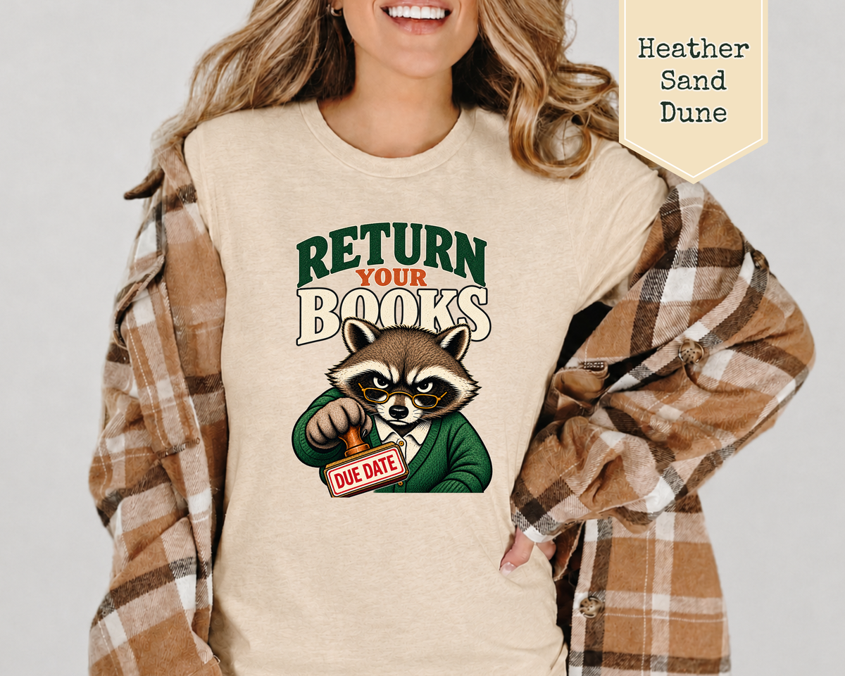 A woman wearing a beige graphic T-shirt with a raccoon holding a 'DUE DATE' sign, layered under a brown plaid flannel shirt. A beige label in the top right reads 'Heather Sand Dune'.